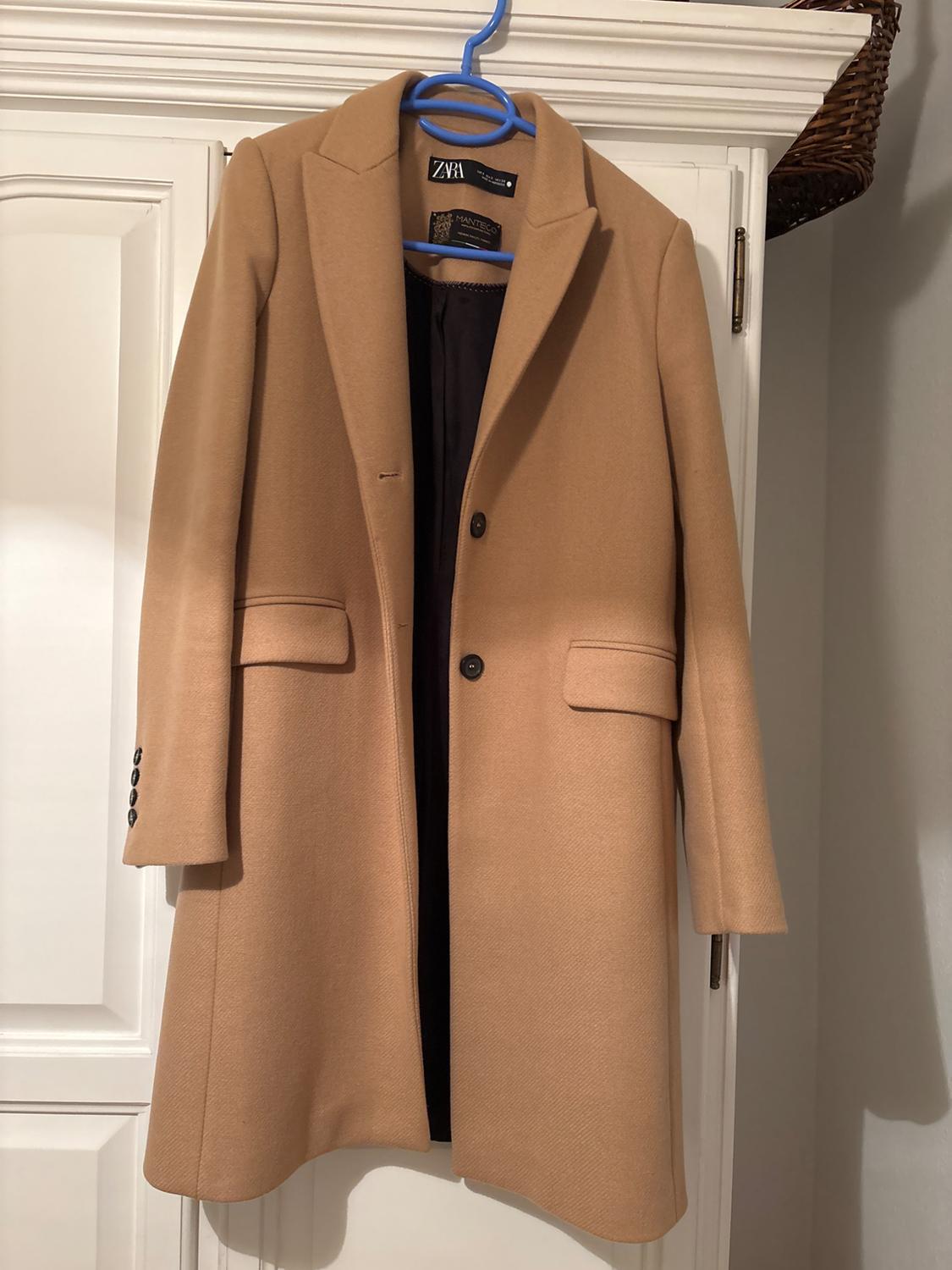 Camel coat hanging on a blue hanger, with lapels and two front pockets. Visible label with the ZARA brand inside.