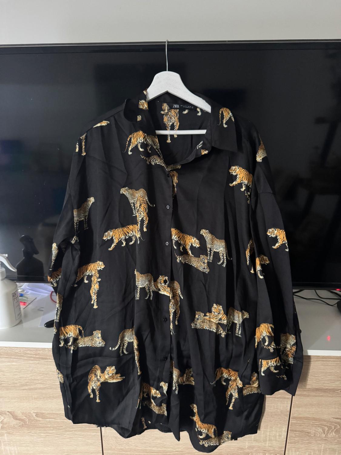 Black long-sleeved shirt with a tiger and leopard print, hanging on a white coat hanger in front of a television.