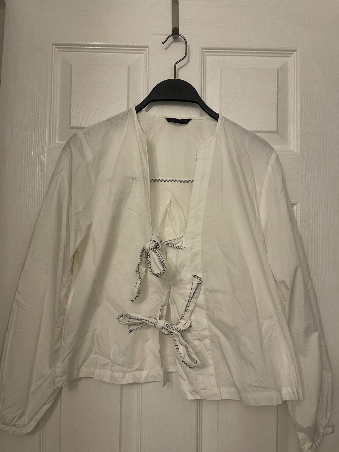 White long-sleeved blouse with ties on the front, hanging on a black hanger over a white door.
