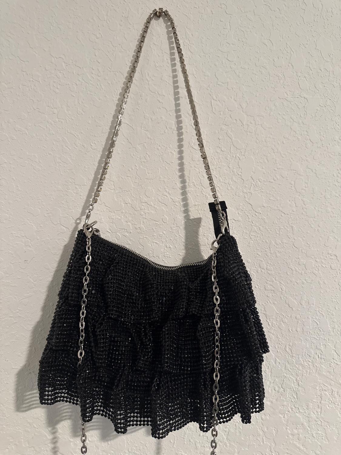 Black bag with mesh texture and ruffles, hanging from a silver chain against a textured white wall.