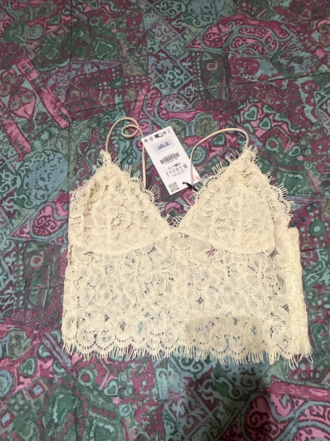 Beige lace top with spaghetti straps on a geometric print bedspread in green and pink tones. Visible tag.