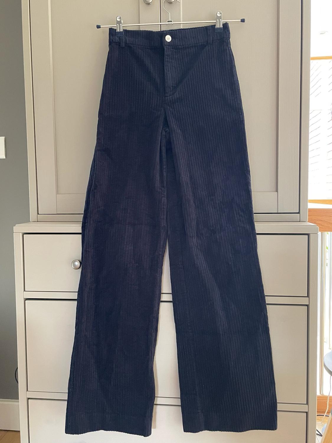 Dark corduroy trousers hanging on a hanger in front of a light wood chest of drawers. Casual style in a homely setting.