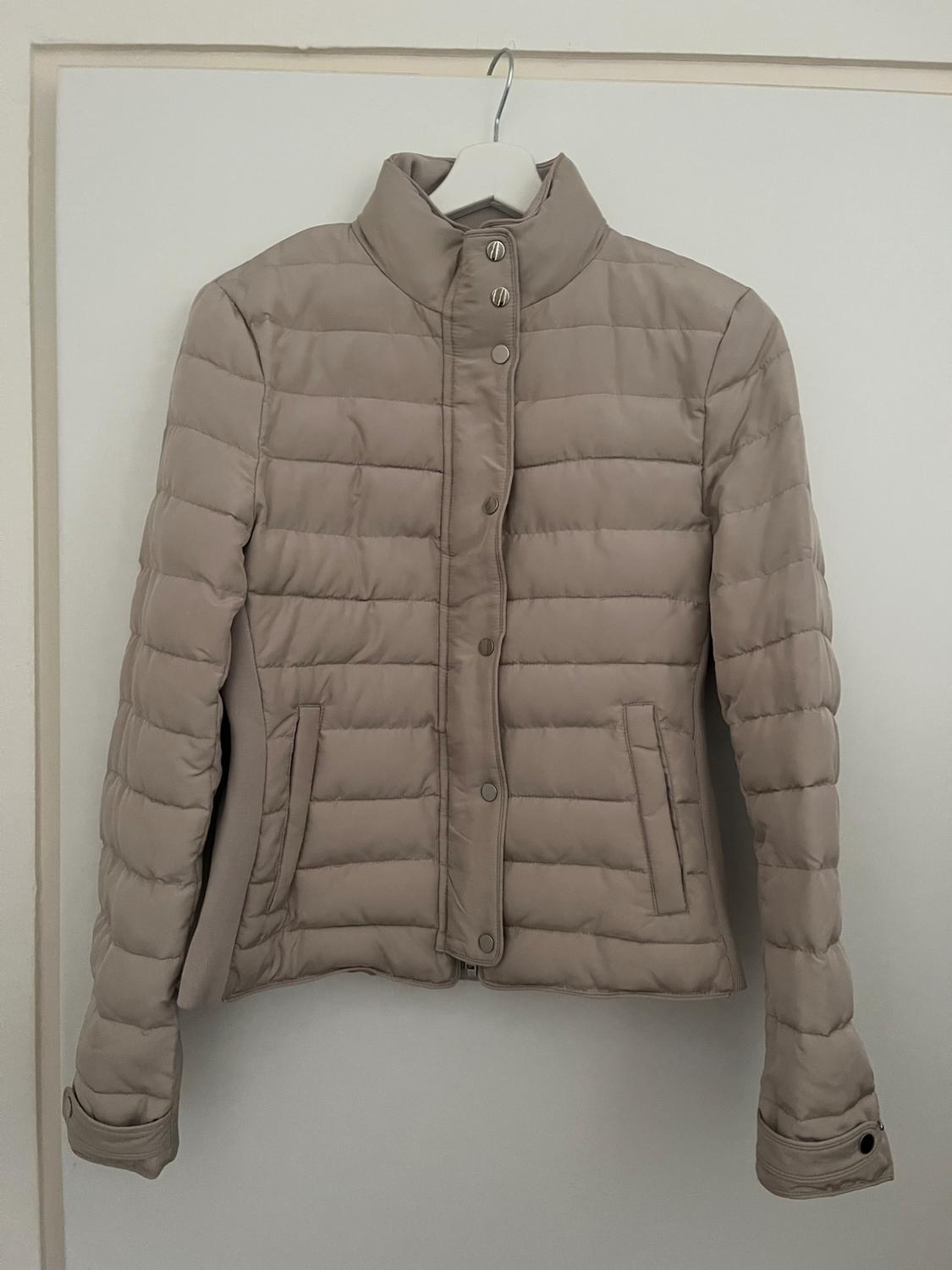 Beige padded jacket hanging on a white hanger. It has button fastening and side pockets. White background.