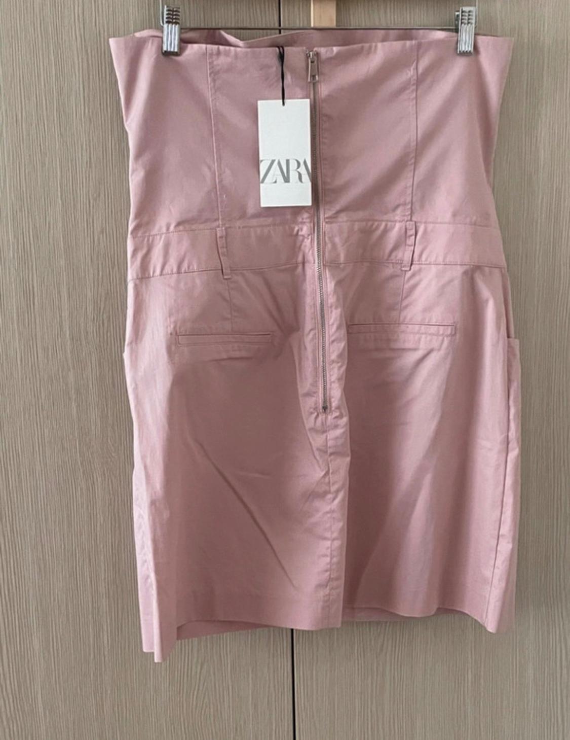 Light pink dress with a front zip and a label bearing the text ZARA, hanging on a hanger against a light wood background.