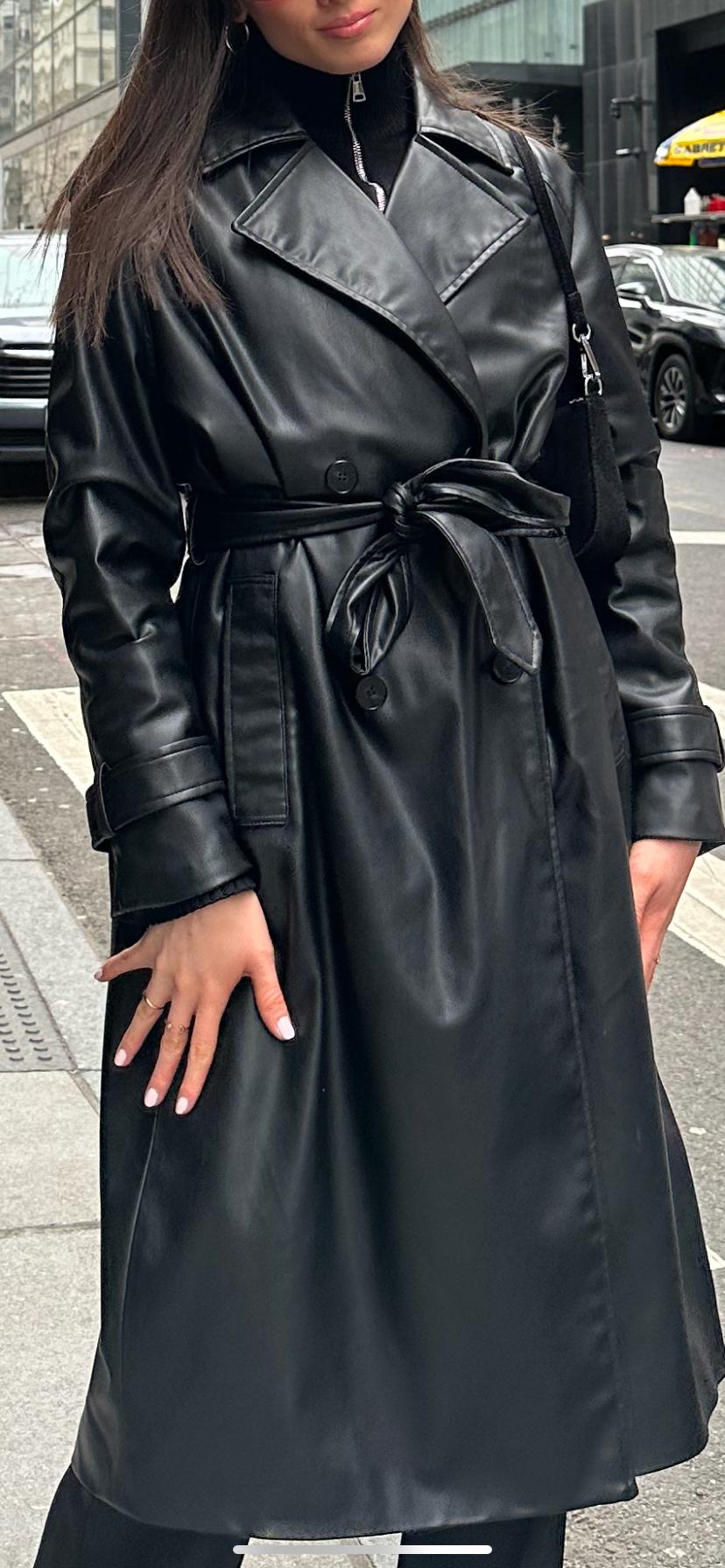 Woman in the street wearing a long black faux-leather coat with a belted waist and a zip-up top. Urban style.
