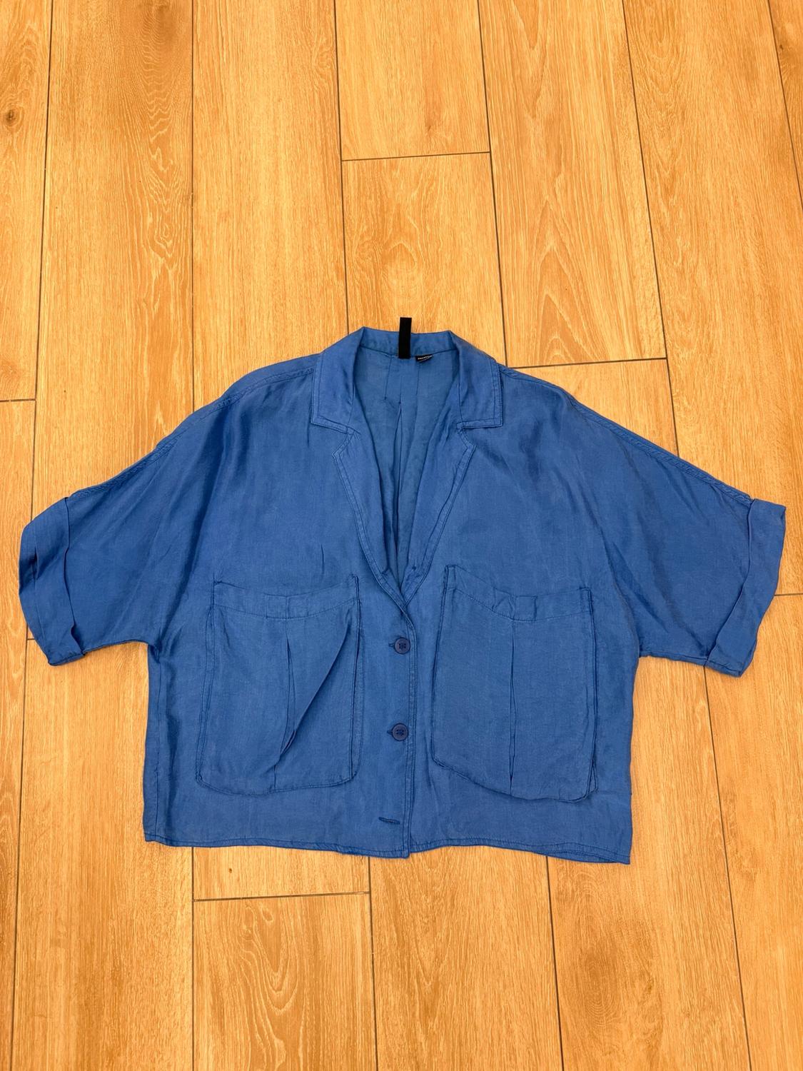 Blue short-sleeved shirt with a collar, buttons and two large front pockets, laid out on a wooden floor.