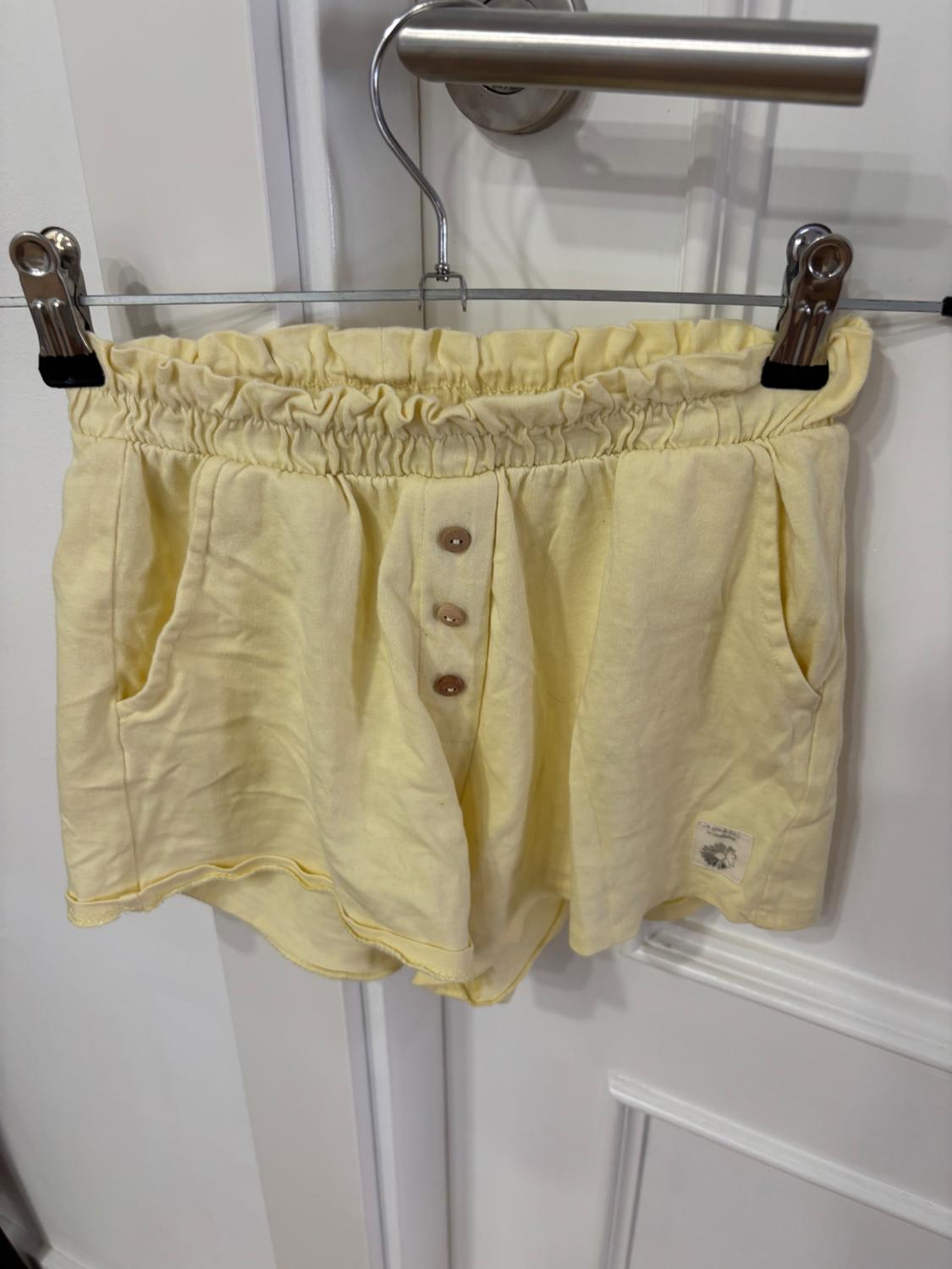 Yellow shorts with an elasticated waistband and buttons on the front, hanging on a metal coat hanger over a white door.