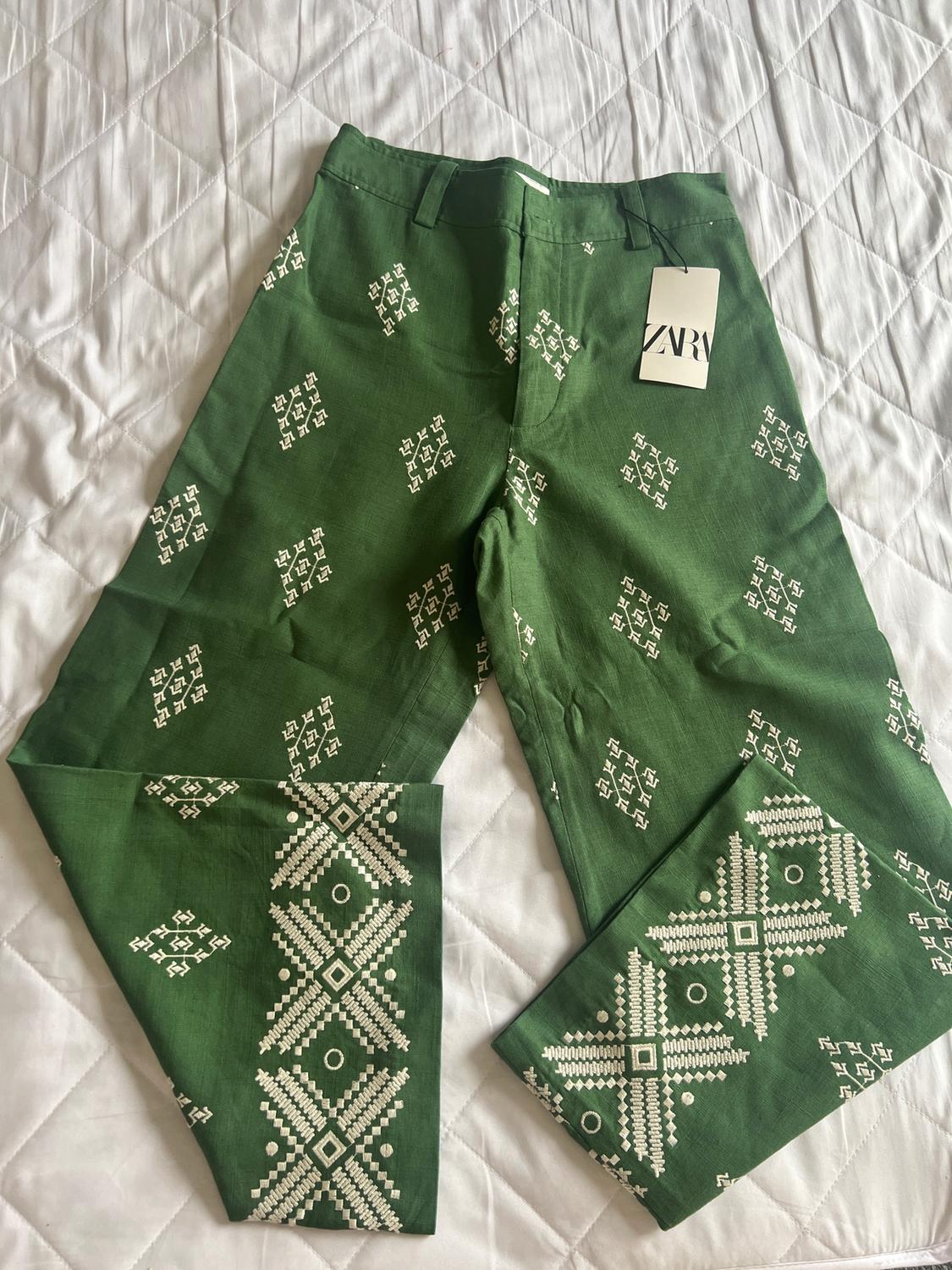 Green trousers with white geometric print on a quilted white bedspread. Visible label with text "ZARA".