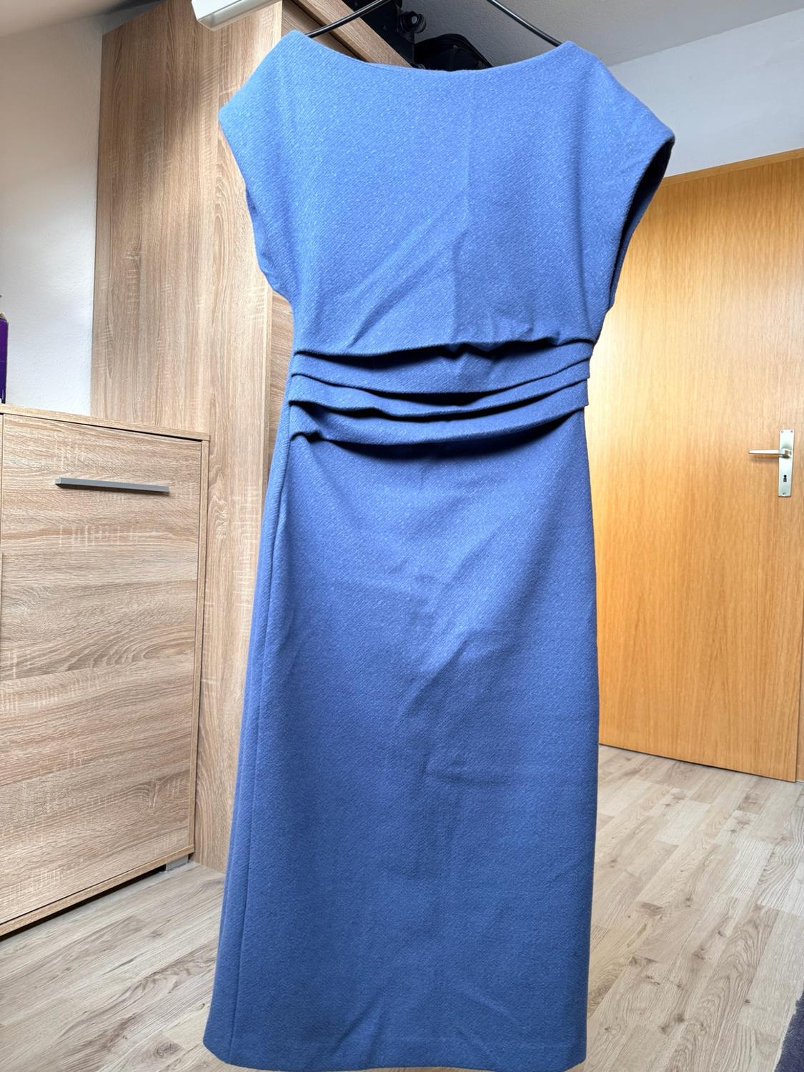 Sleeveless blue dress with pleats at the waist, hanging on a hanger in a room with a wardrobe and wooden door.