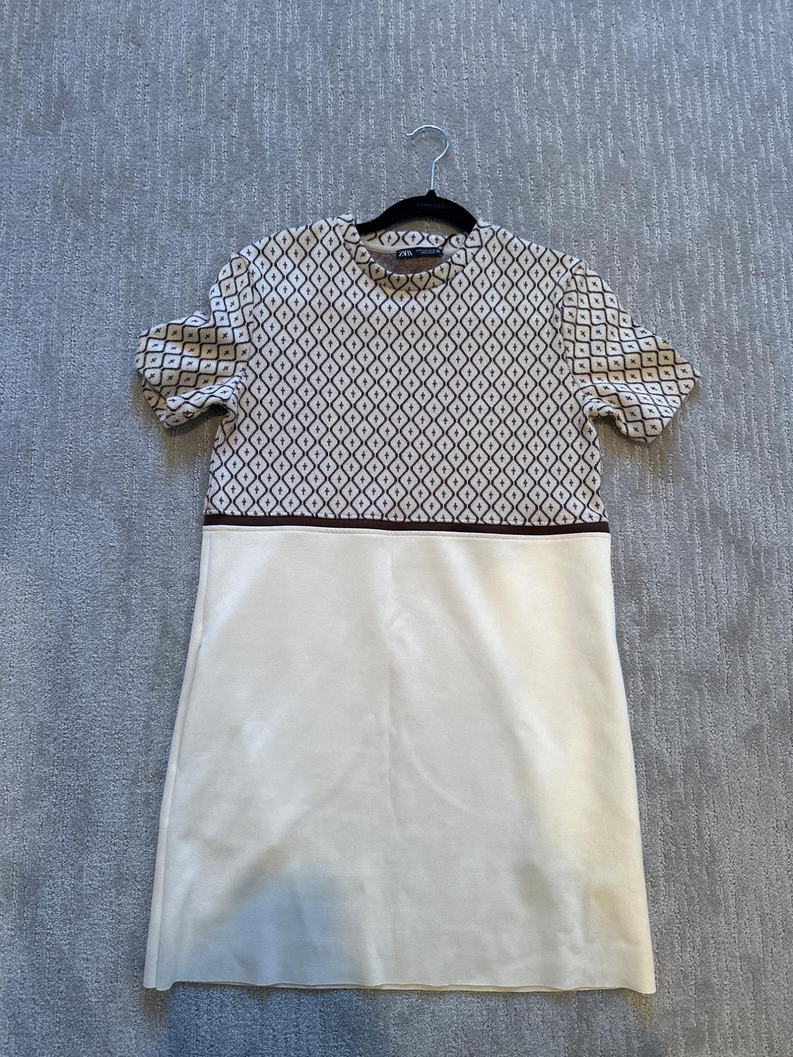 Short-sleeved dress with geometric print on the top and a plain white skirt, hanging on a hanger over a gray carpet.