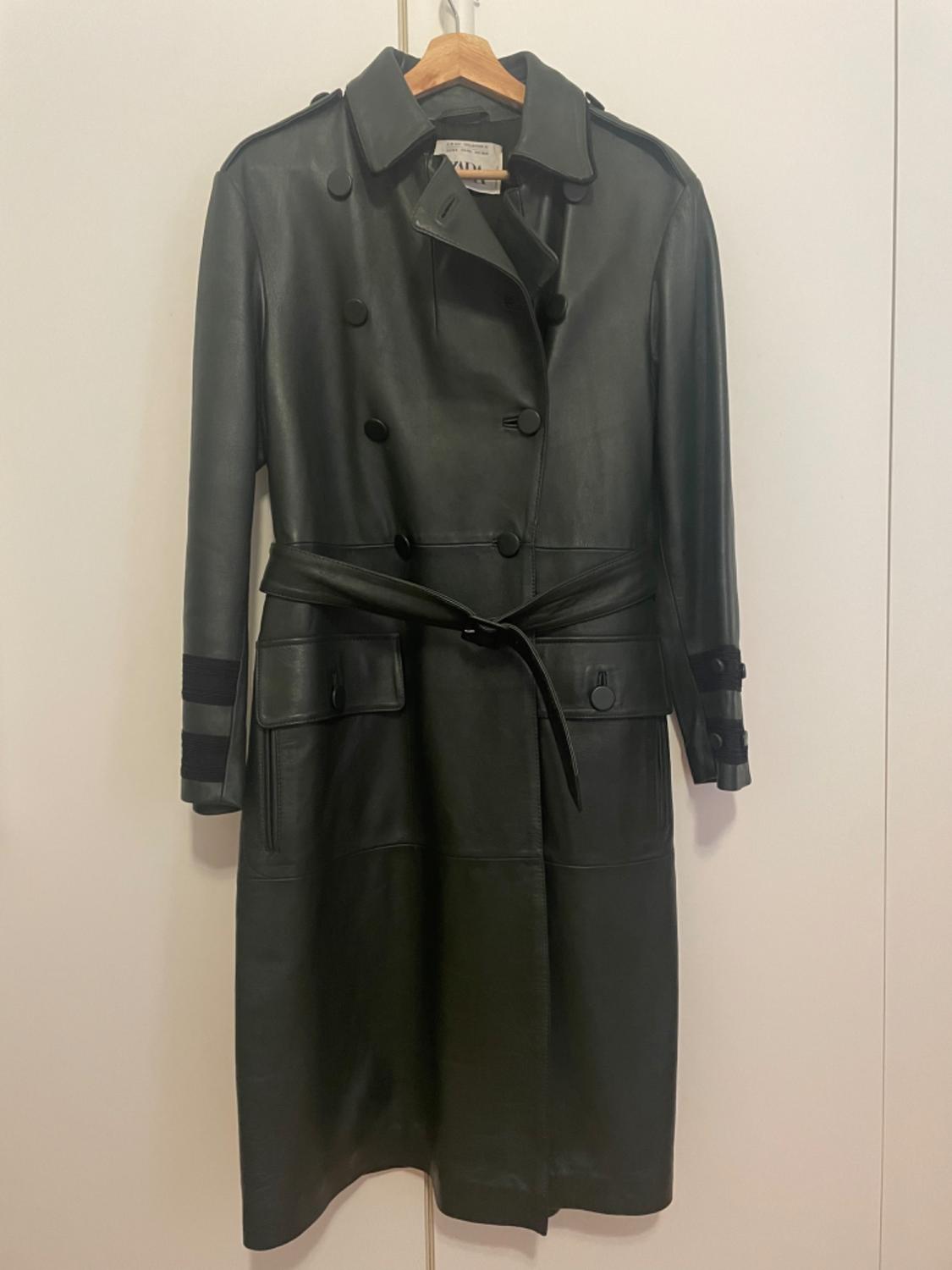 Black leather coat hanging on a wooden hanger, double-breasted with a belt, against a white background.