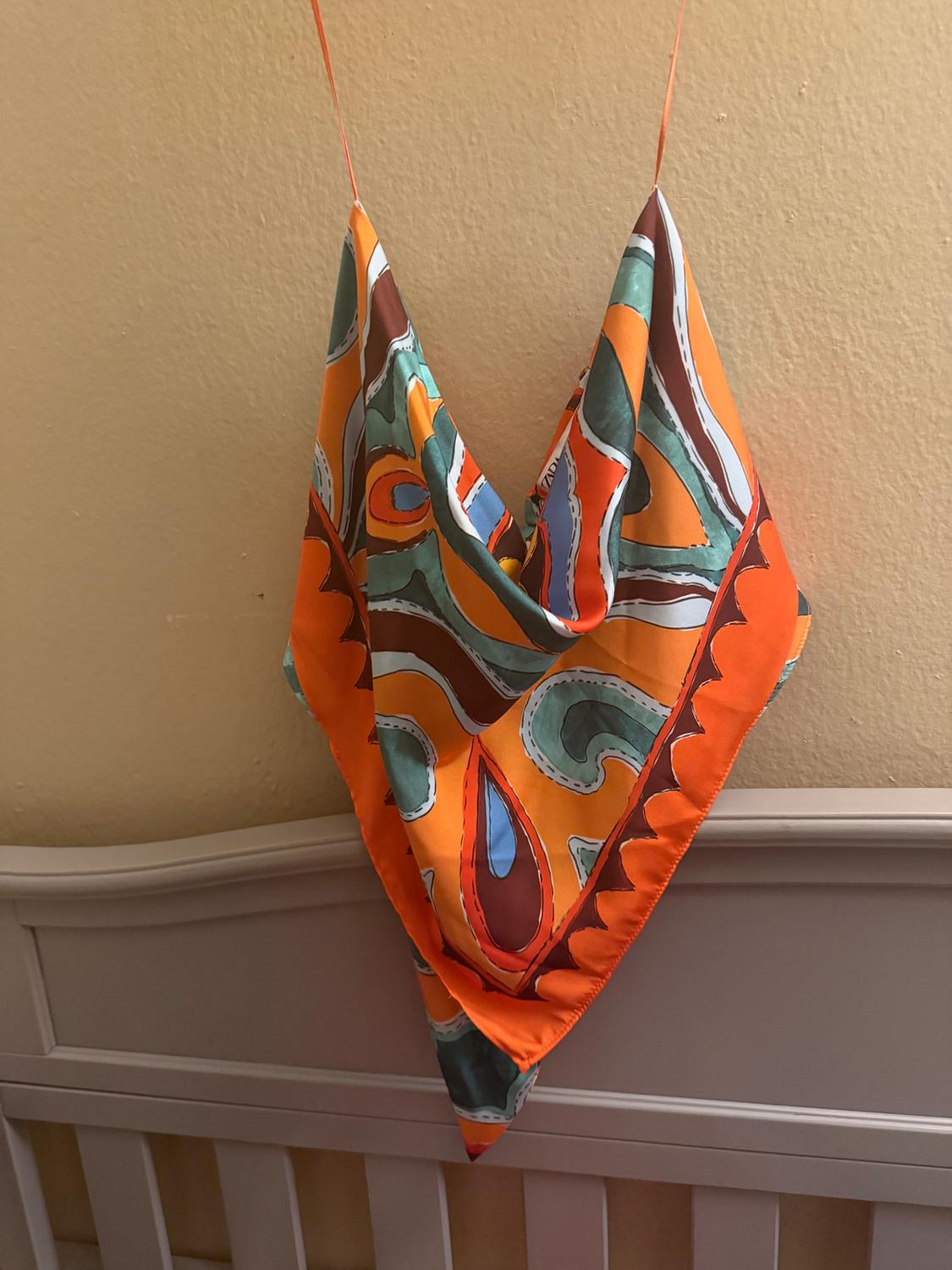 Hanging scarf with a colorful print in orange, green, and blue tones, against a beige wall background and a bed headboard.