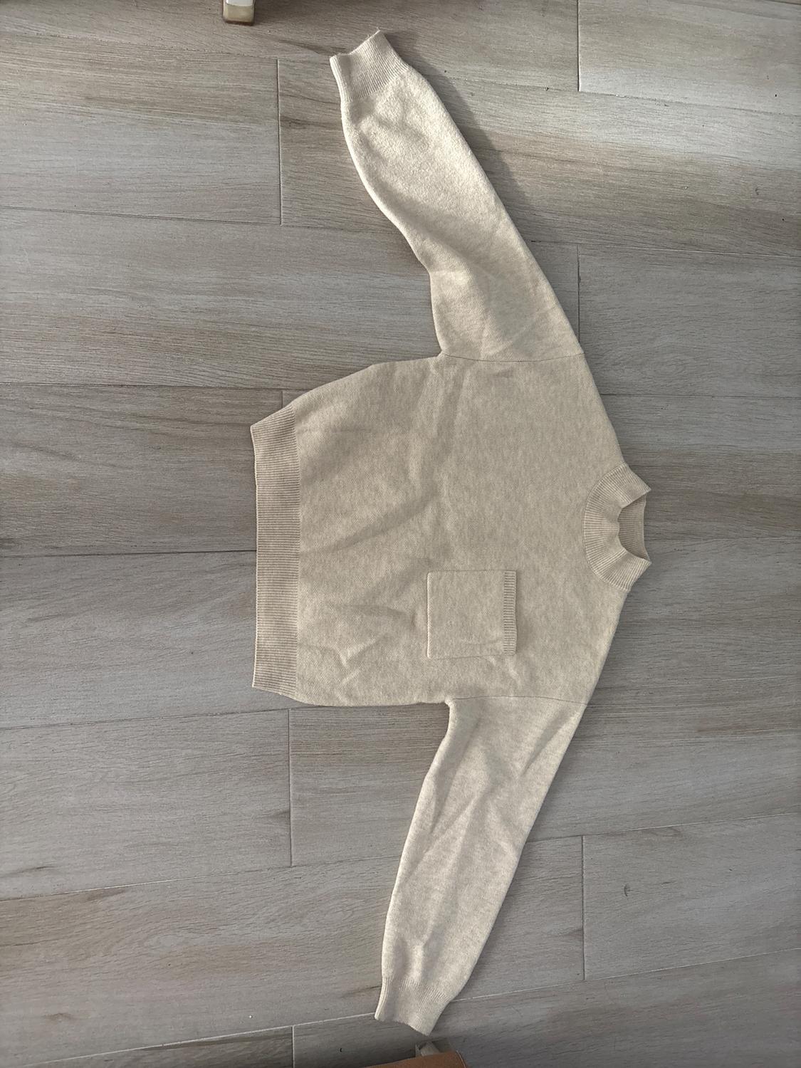 Beige long-sleeved jumper with a front pocket, laid out on a light wooden floor.