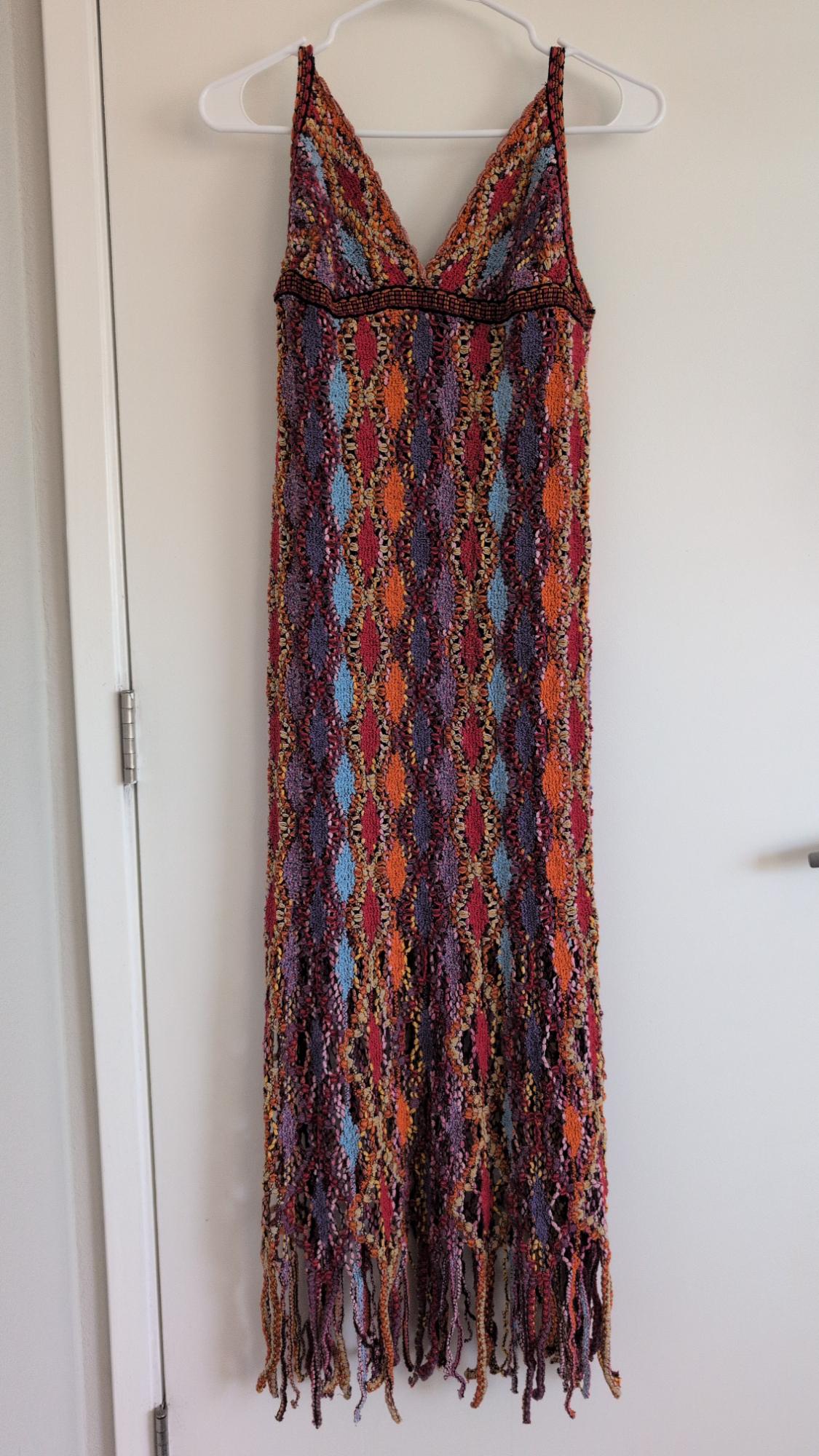 Long-sleeved V-neck knit dress with a multicolored argyle pattern and a fringed hem, hanging on a hanger against a white door.