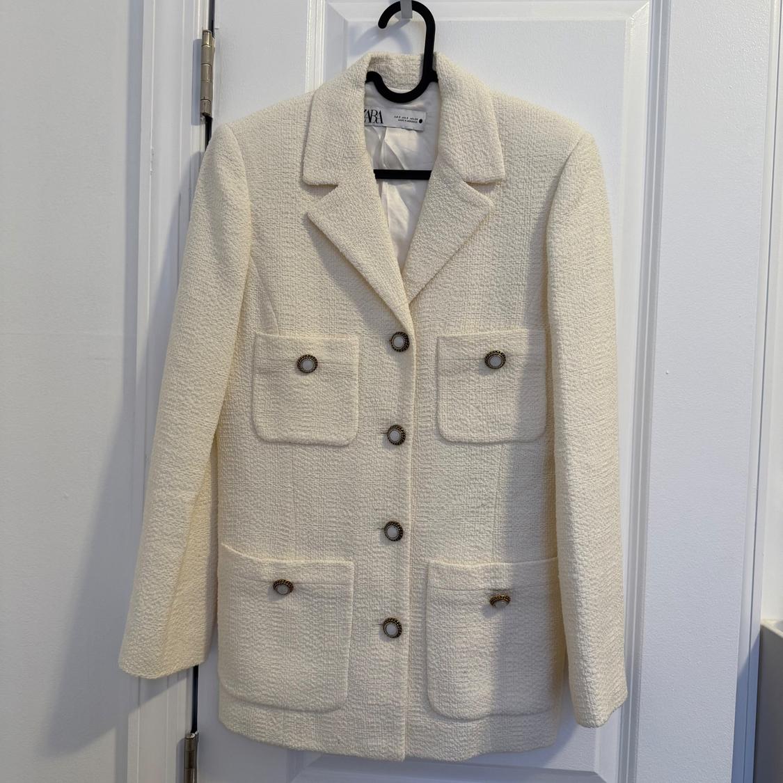 White wool jacket hanging on a black hanger over a white door. It has four front pockets with buttons.