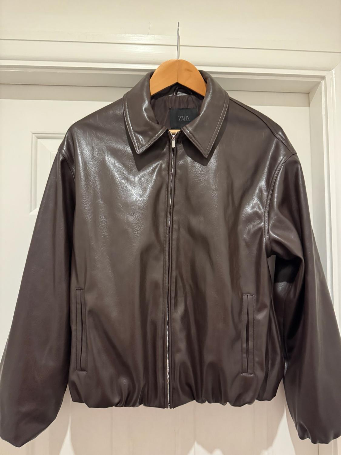 Dark brown jacket with a front zip, hanging on a wooden hanger over a white door. Inner label with the ZARA text.