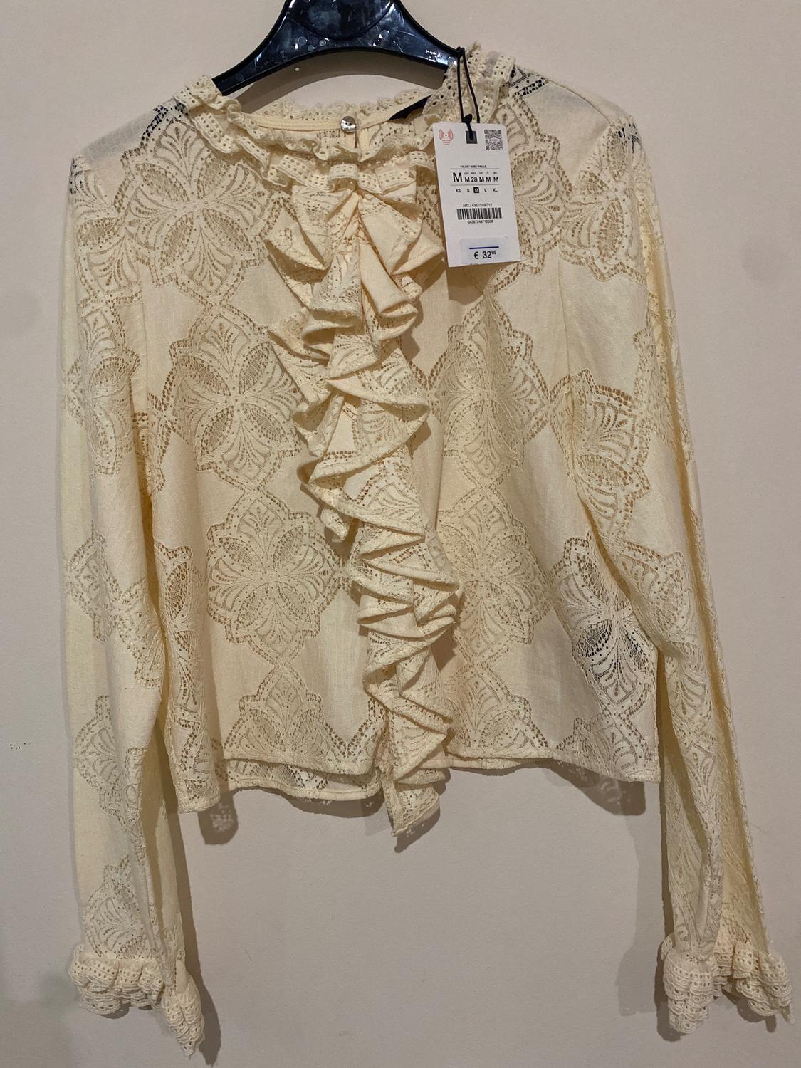 Beige lace blouse with front ruffles, hanging on a black hanger. Visible tag with price of €32.