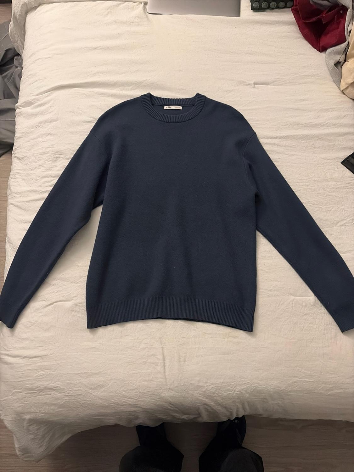 Navy blue crew neck sweater on white sheets. ZARA label. A person's feet are visible at the bottom.