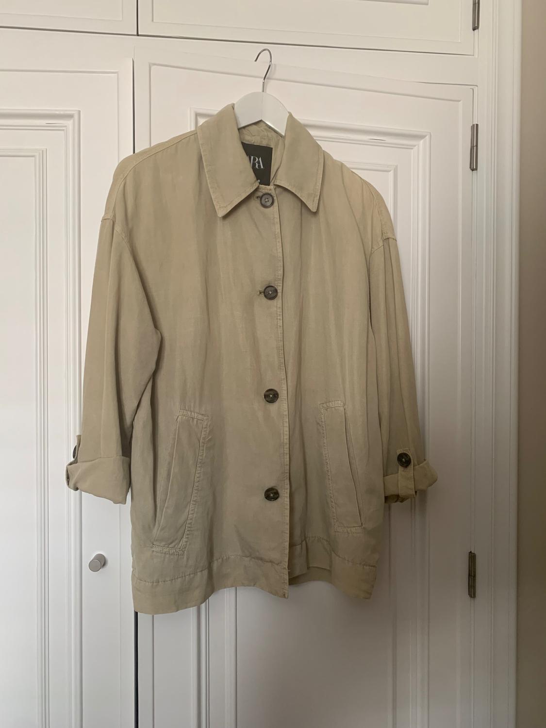 Beige jacket hanging on a white coat hanger in front of a white door. The jacket has large buttons and front pockets.