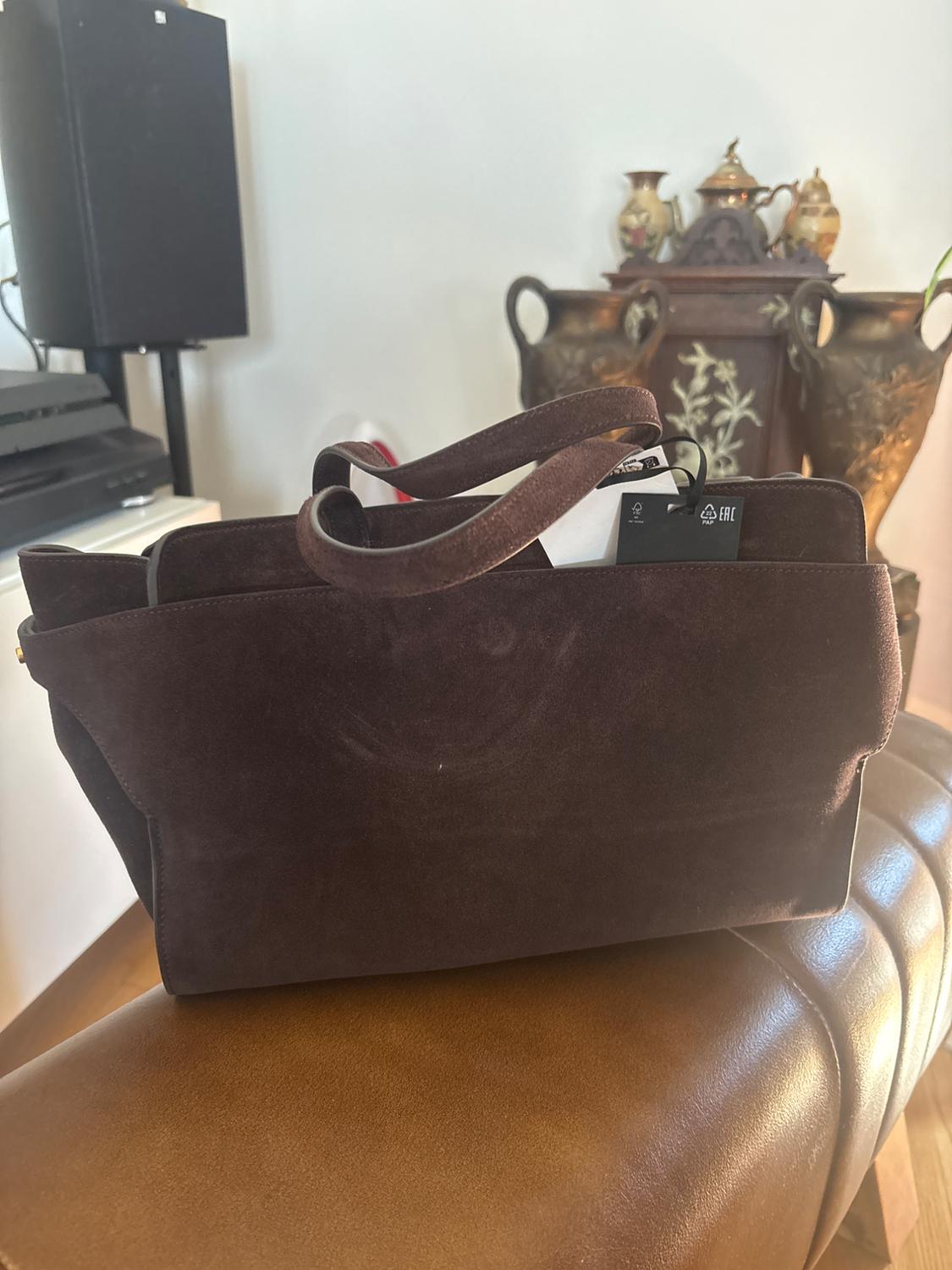 Dark brown suede bag with EAC labels and text, on a leather seat in a living room with furniture and decor in the background.
