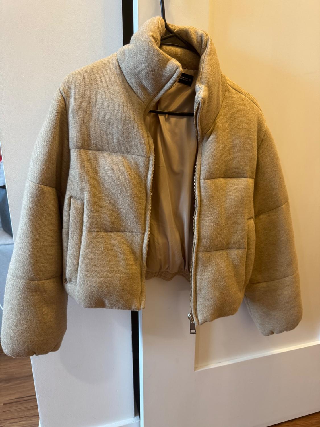 Beige padded jacket with a high collar and open zip, hanging from a black hanger on a white door.