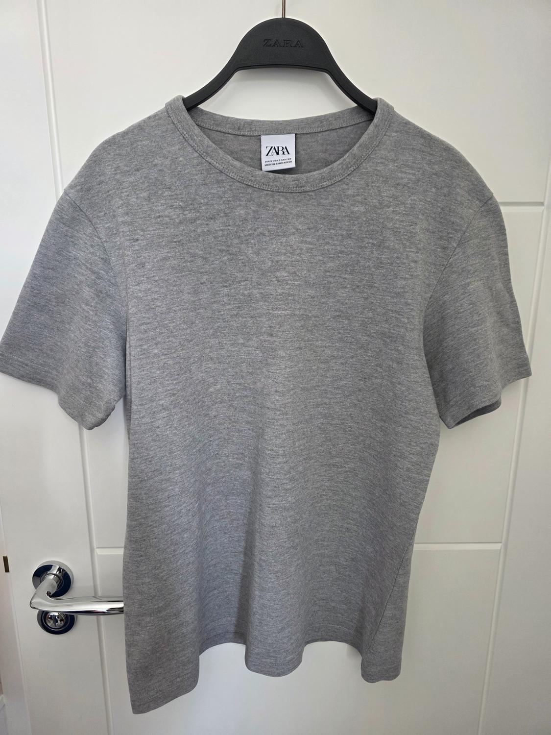 Grey short-sleeved T-shirt hanging on a black hanger over a white door. Visible text: ZARA.