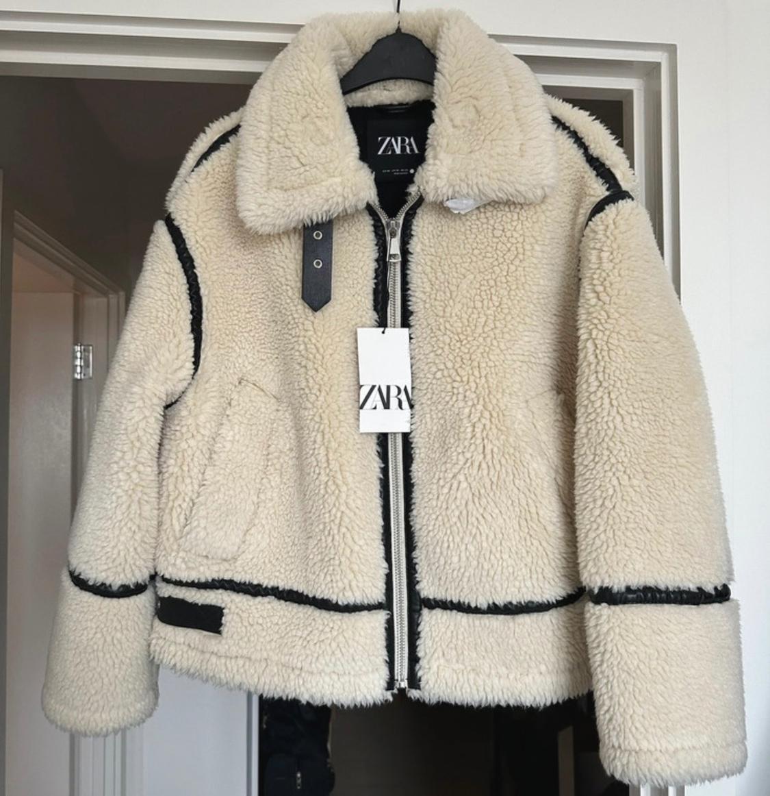 Cream-coloured jacket with a curly texture, black trims and a zip, hanging on a hanger. Includes labels bearing the text ZARA.