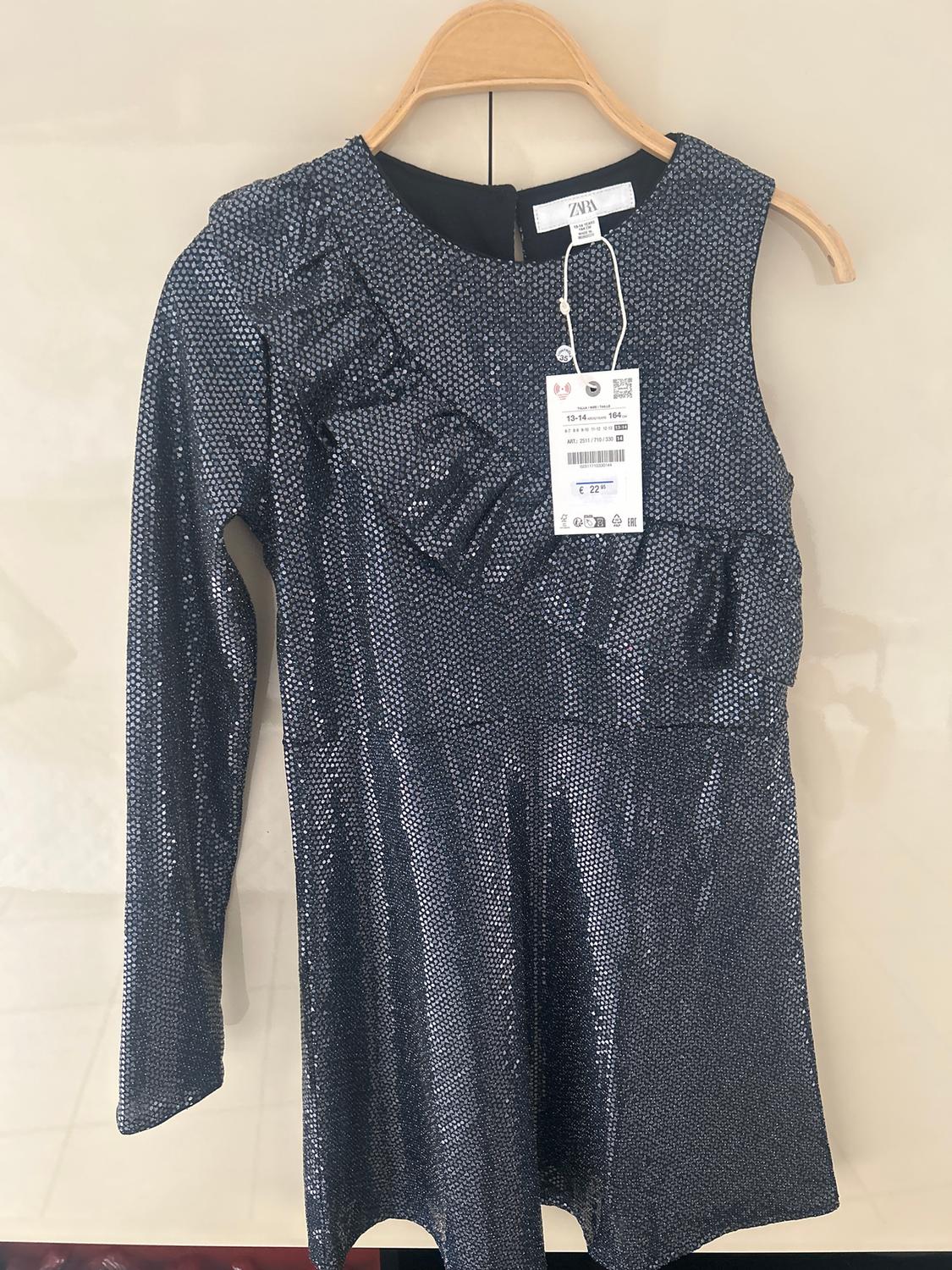 Dark blue sparkly dress with frills on the bust, long sleeves, hanging on a wooden hanger. Visible tag with a price of €22.