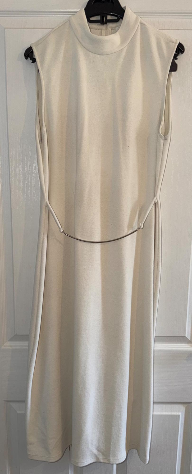 Cream sleeveless dress with a high neck and a thin belt, hanging on a black hanger in front of a white door.