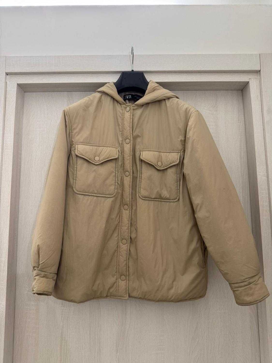 Beige padded jacket with a hood, buttons and front pockets, hanging on a coat hanger over a wooden door. ZARA label.