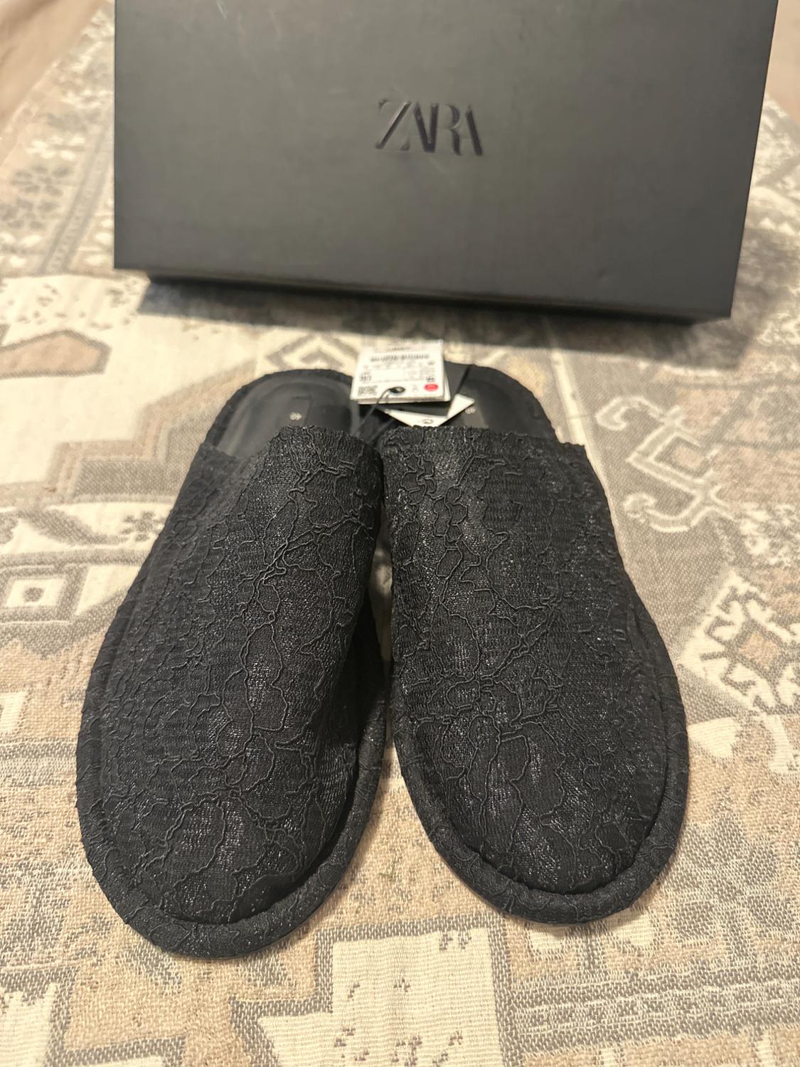 Black trainers with a lace texture on a patterned rug. Black box with "ZARA" logo in the background. Label visible on the trainers.