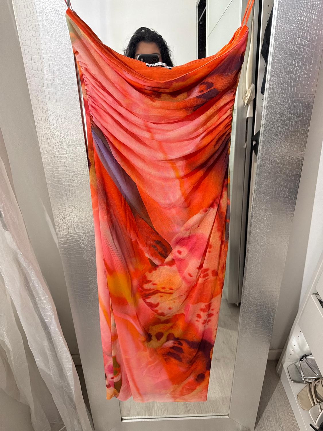 Long dress with an abstract print in red and orange tones, held by a person in front of a mirror in a changing room.