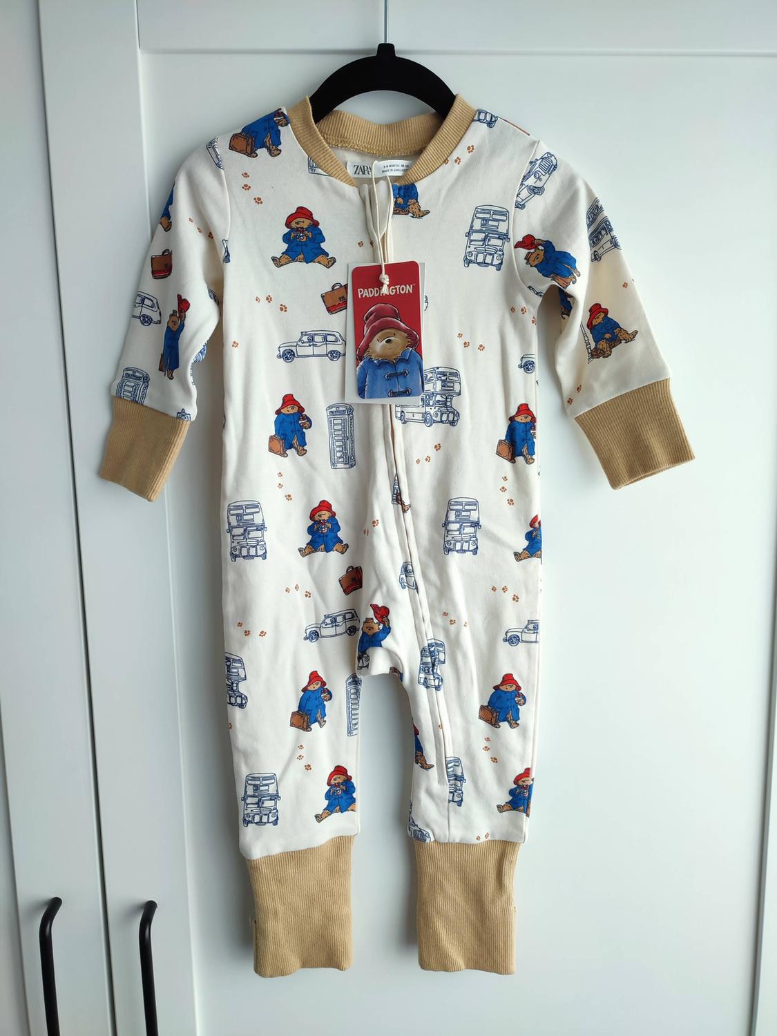 Cream baby pyjamas with a Paddington print and buses. Mustard cuffs. Hung on a hanger. Labels with the text PADDINGTON and ZARA.