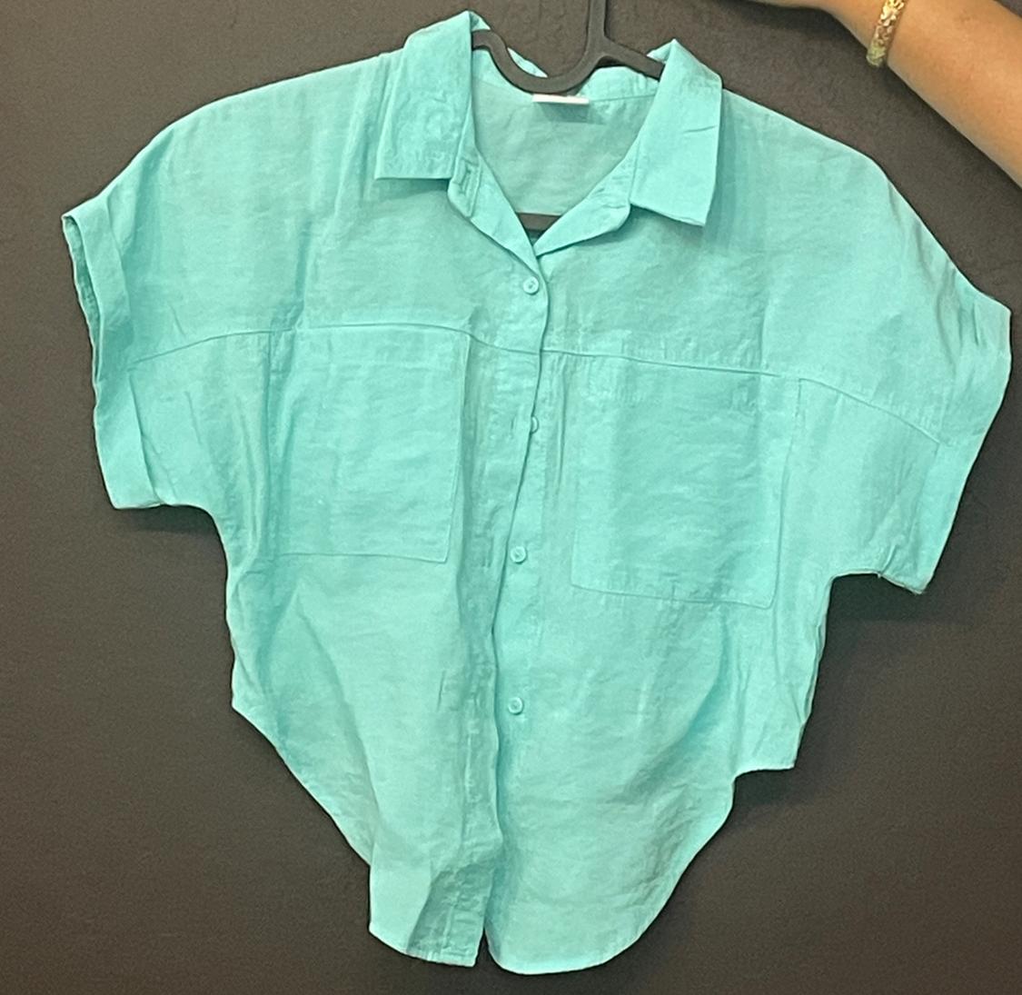 Turquoise short-sleeved button-down shirt with pockets, hanging on a hanger held by an arm with a bracelet against a dark background.