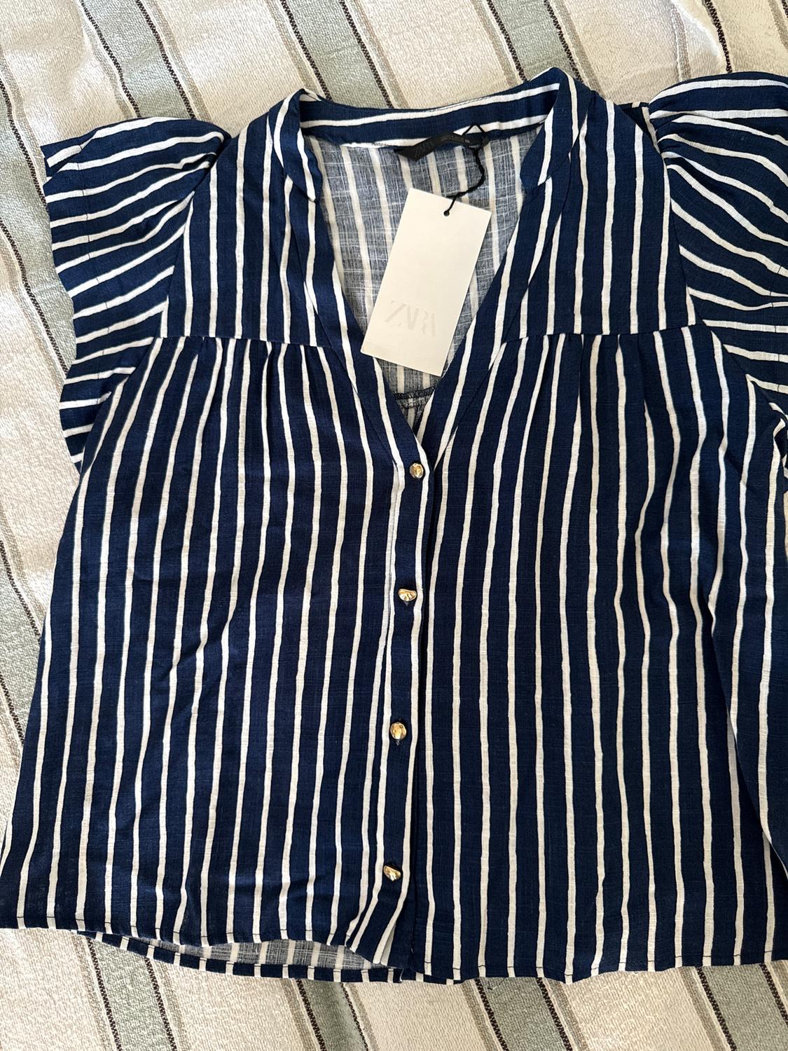 Blue and white striped blouse with gold buttons on a plain background fabric. White hanging tag with "ZARA" text.