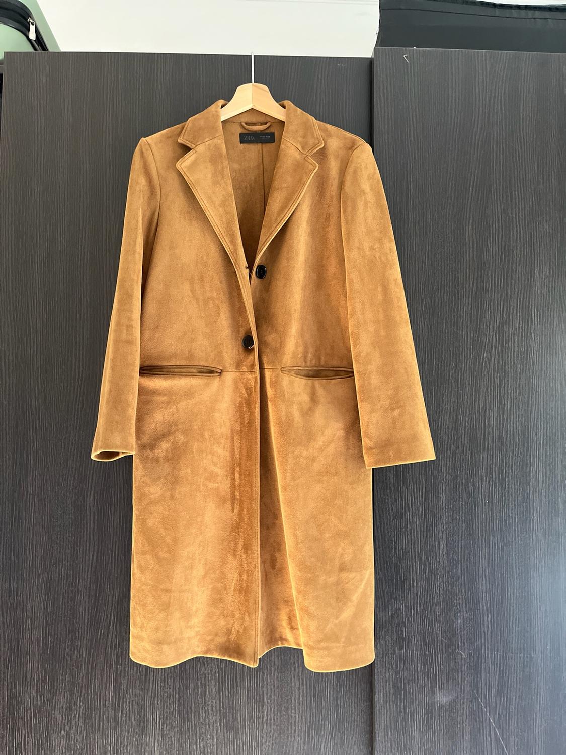 Long camel-coloured coat with a soft texture, lapels and two buttons, hanging on a wooden hanger against a dark background. Text: ZARA.