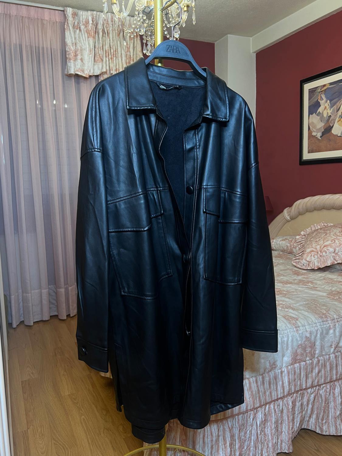 Black leather coat hanging on a Zara hanger, in a bedroom with pink curtains and a bed with a patterned bedspread.