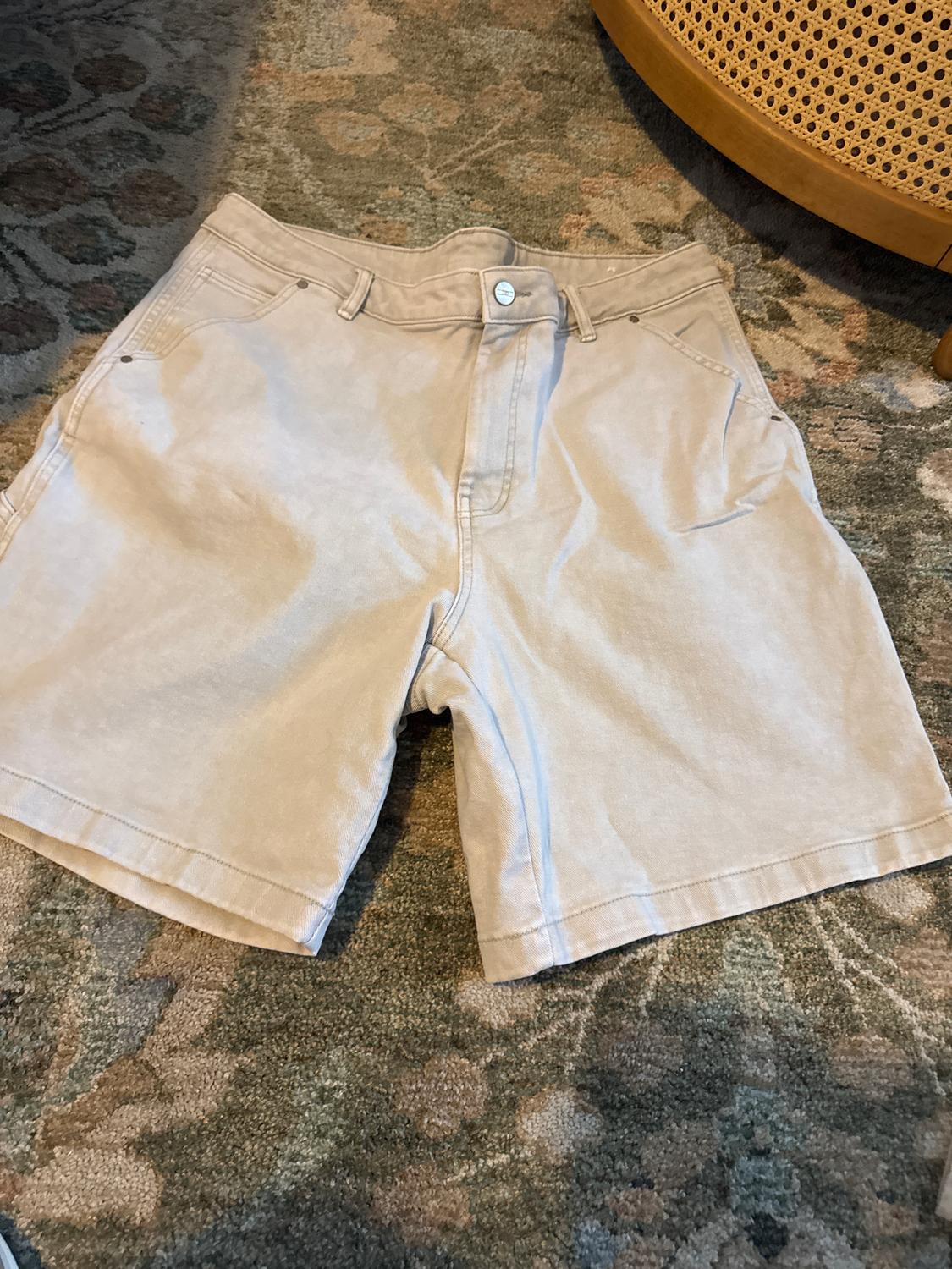 Beige shorts on a rug with a floral design. Visible metal button on the waistband. Relaxed and homely atmosphere.