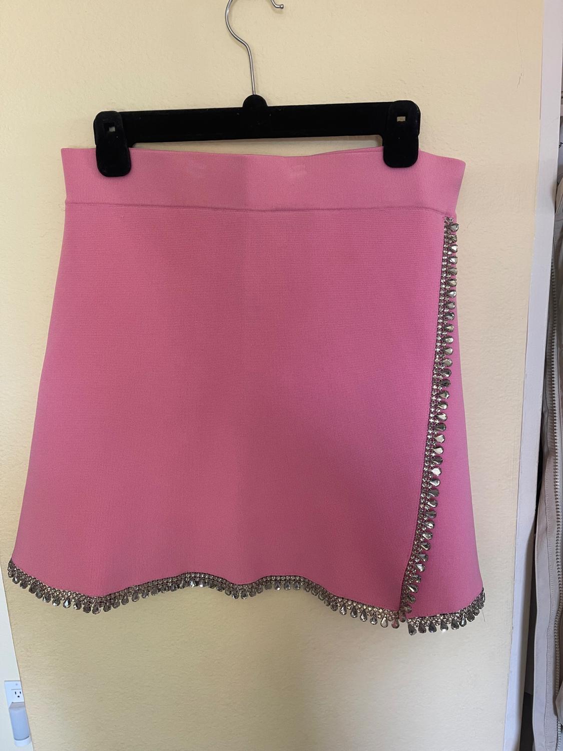 Pink mini skirt with silver rhinestone-decorated edges, hanging on a black hanger against a beige wall.