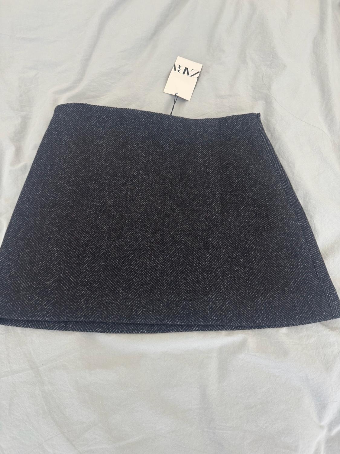 Dark grey herringbone miniskirt on a crumpled light-coloured fabric. It has a label bearing the text ZARA.