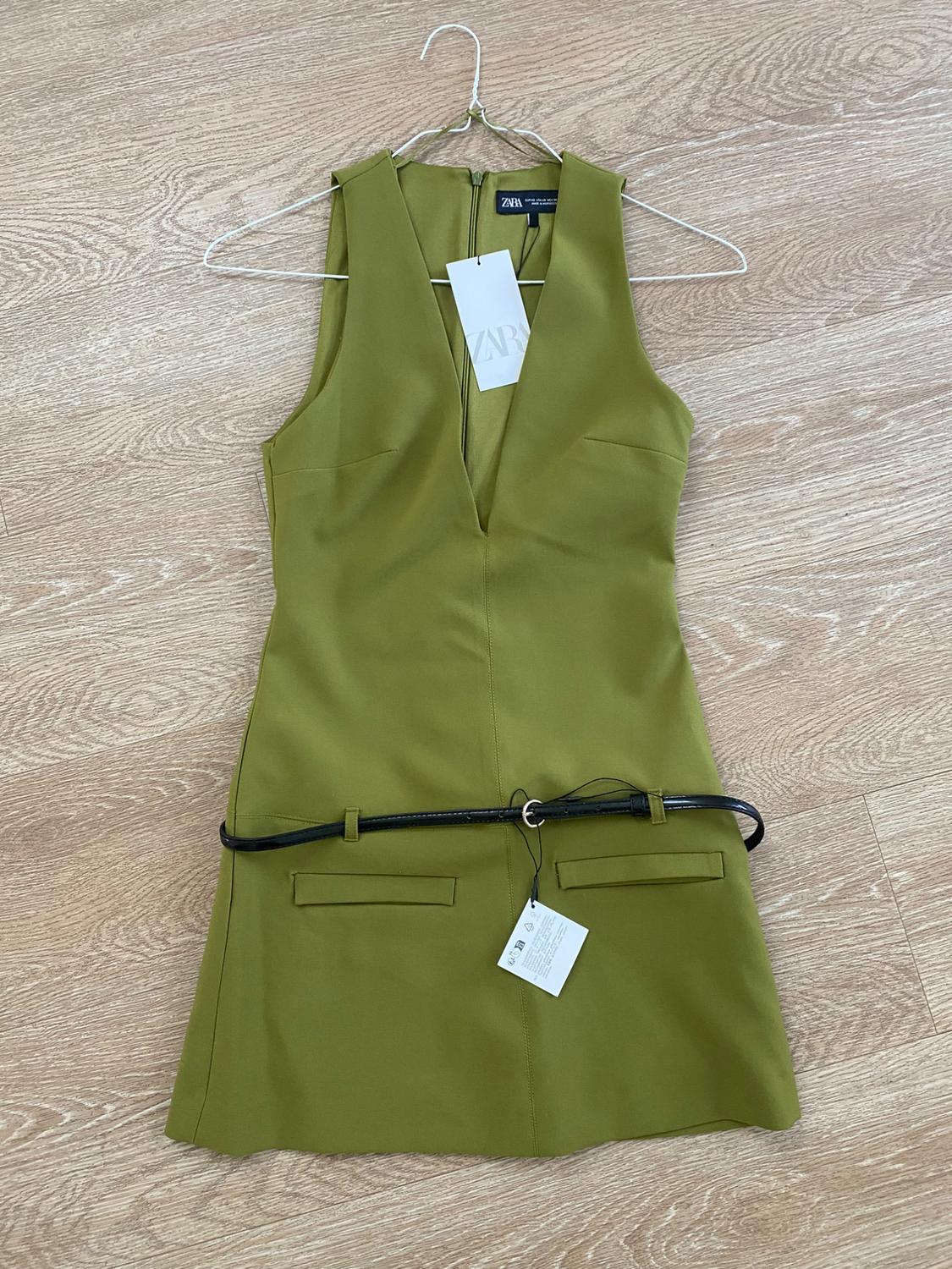 Sleeveless olive green V-neck dress with black belt and hangtags, on a light wood floor.