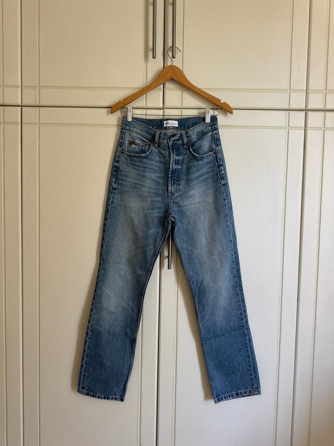 Blue denim trousers hanging on a wooden hanger in front of a white wardrobe. Washed-effect design with an inner label bearing the text ZARA.