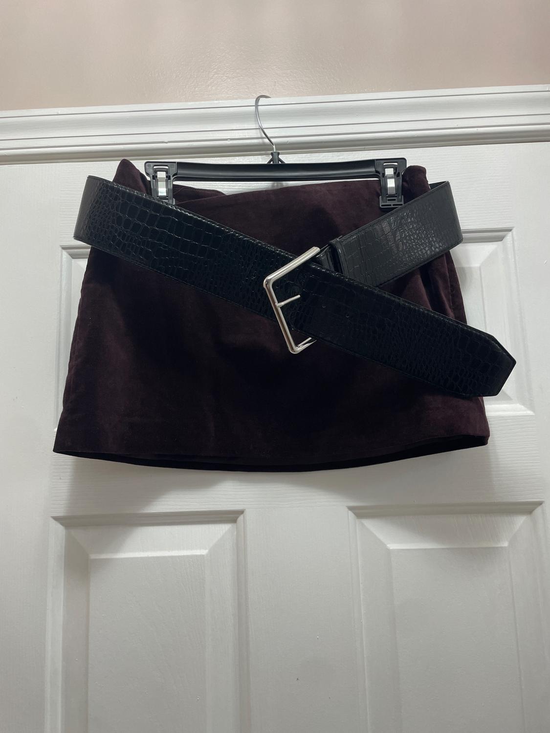 Burgundy miniskirt hanging on a white door. A wide black
belt with a crocodile texture and silver buckle crosses over it.