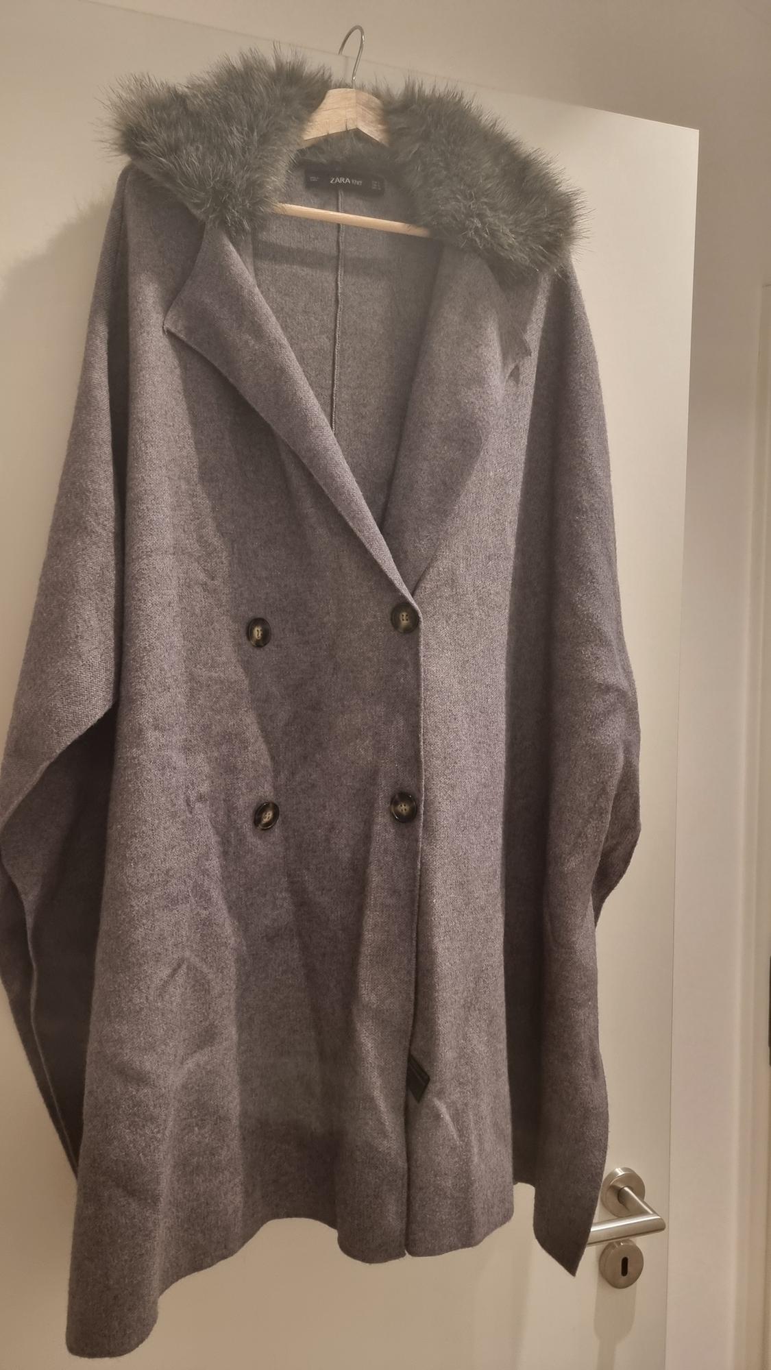 Grey coat with black buttons and a faux fur collar hanging on a white door. Visible label: "ZARA".