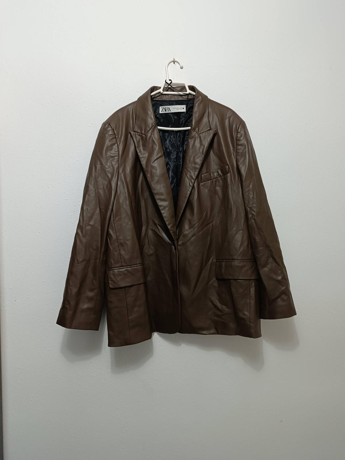 Brown jacket hanging on a white hanger against a white wall. Visible label with the text "ZARA".