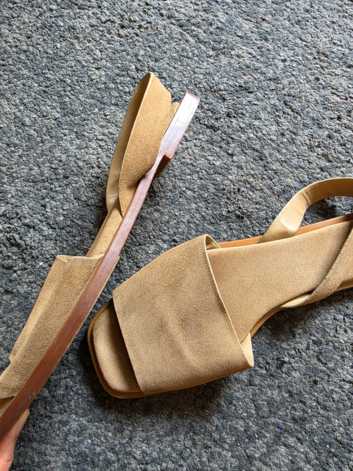 Camel-coloured suede flat sandals with a wide strap, arranged casually on a textured grey rug.