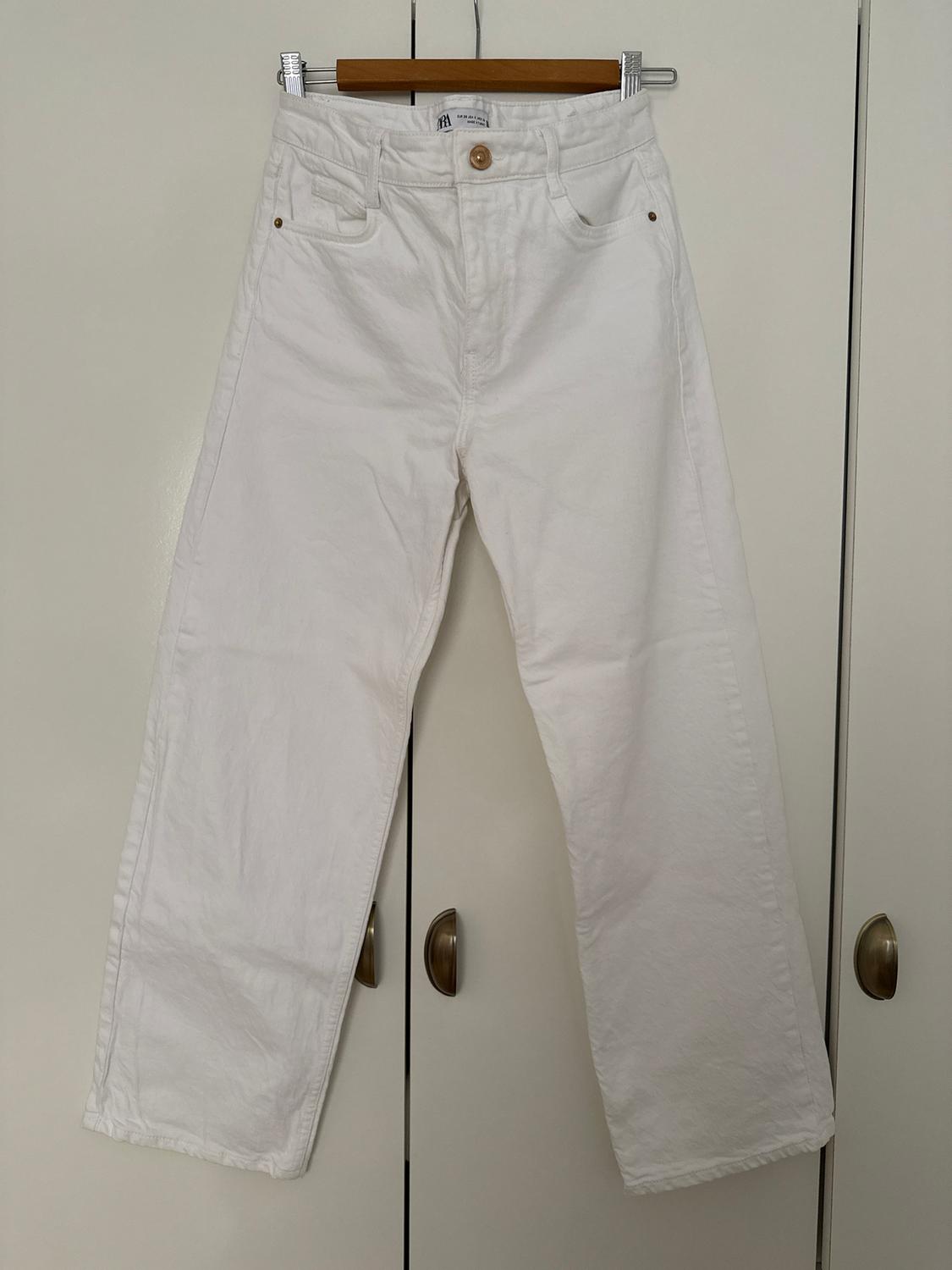 White wide-leg trousers hanging on a wooden hanger in front of a white wardrobe with gold handles.