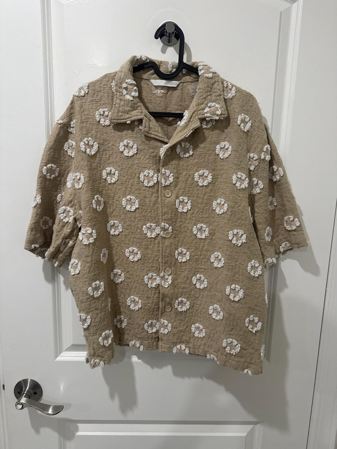 Beige short-sleeve shirt with texture and raised white circular patterns, hanging on a black hanger over a white door.