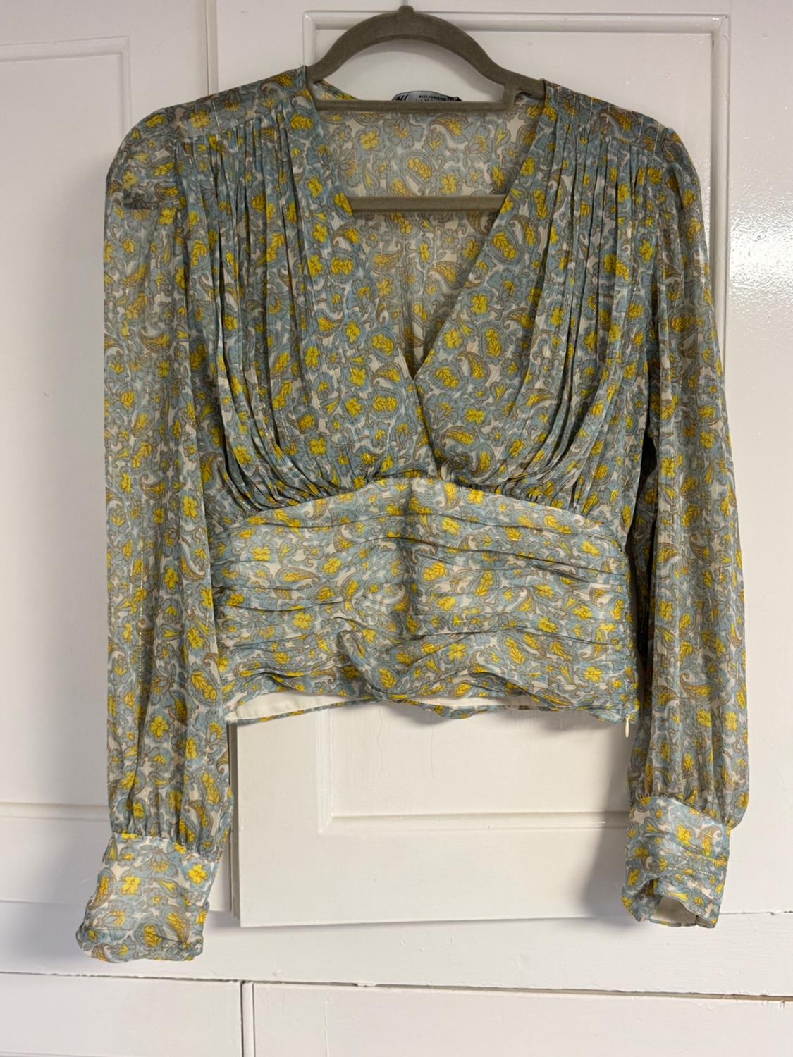 Long-sleeved blouse with yellow and grey floral print on a light blue background, crossover design at the chest and pleats, hanging on a coat hanger.