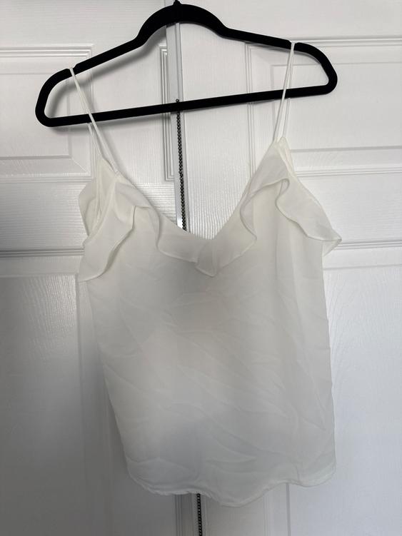 White camisole with ruffles on the neckline, hanging on a black hanger over a white door.